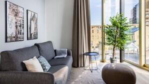 Un salon moderne avec un canapé gris, un pouf rond et une petite table noire. Deux tableaux encadrés ornent le mur, ainsi qu'une plante en pot. De grandes fenêtres offrent une vue sur les bâtiments environnants.