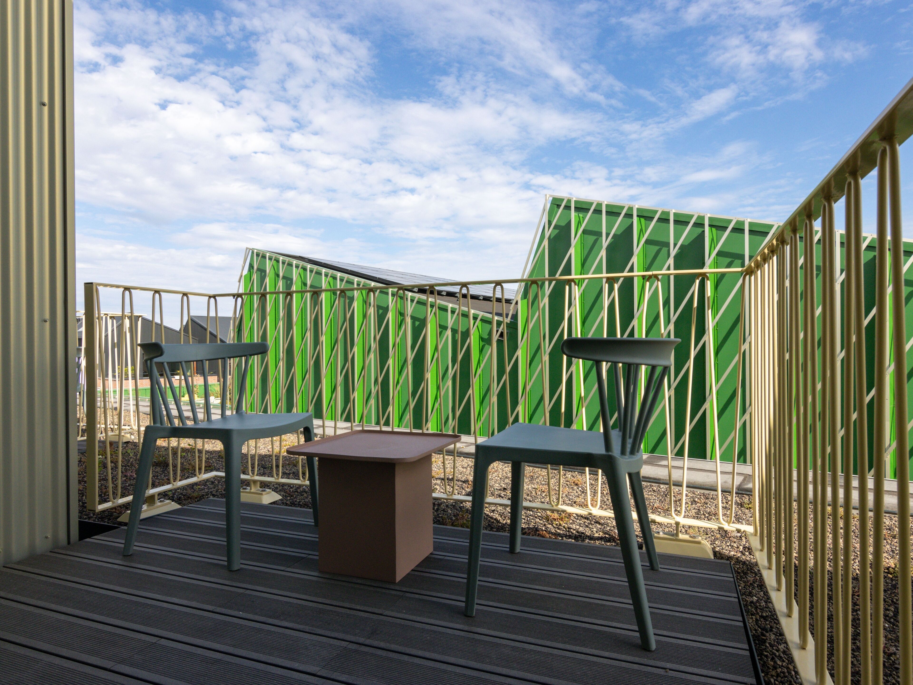 Een terras met twee stoelen en een tafel, omgeven door een groene hek en een metalen leuning.