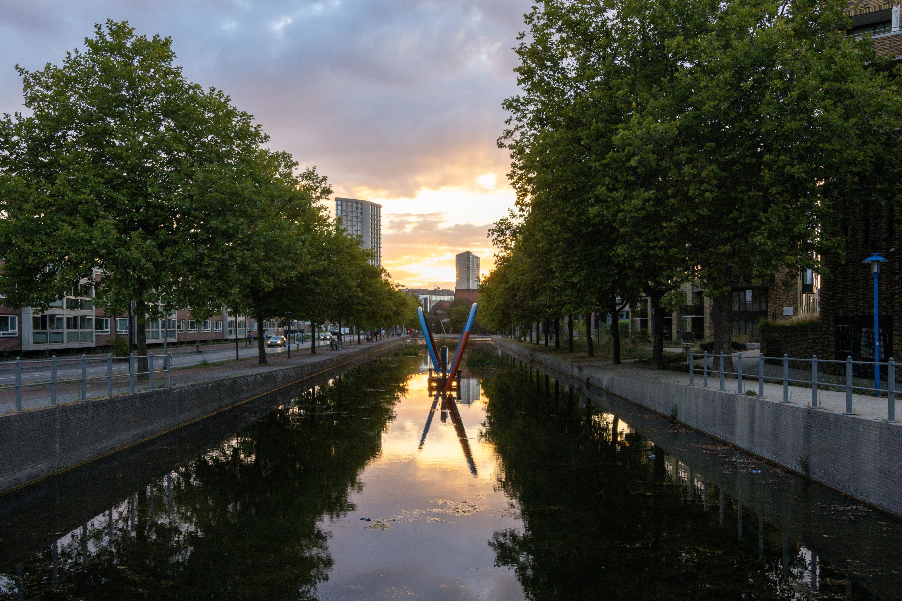 A canal with a bridge, trees, and buildings in the background.