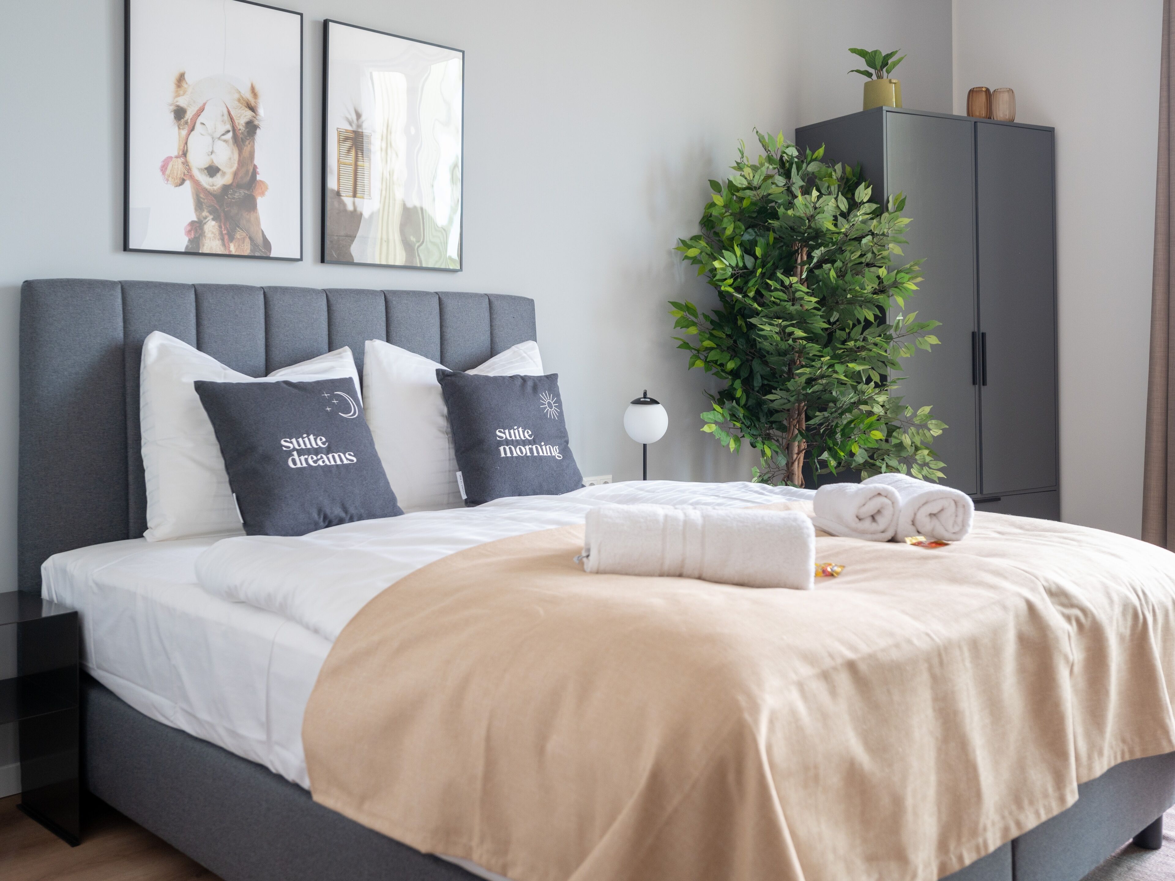 Une chambre moderne avec une tête de lit rembourrée de couleur grise, un grand lit avec une literie blanche et beige, deux coussins ornés de motifs, une plante en pot et une armoire grise.