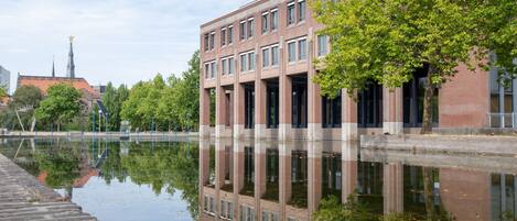 Un bâtiment à la façade en briques et aux grandes fenêtres, dont le reflet se mire dans une étendue d’eau calme.