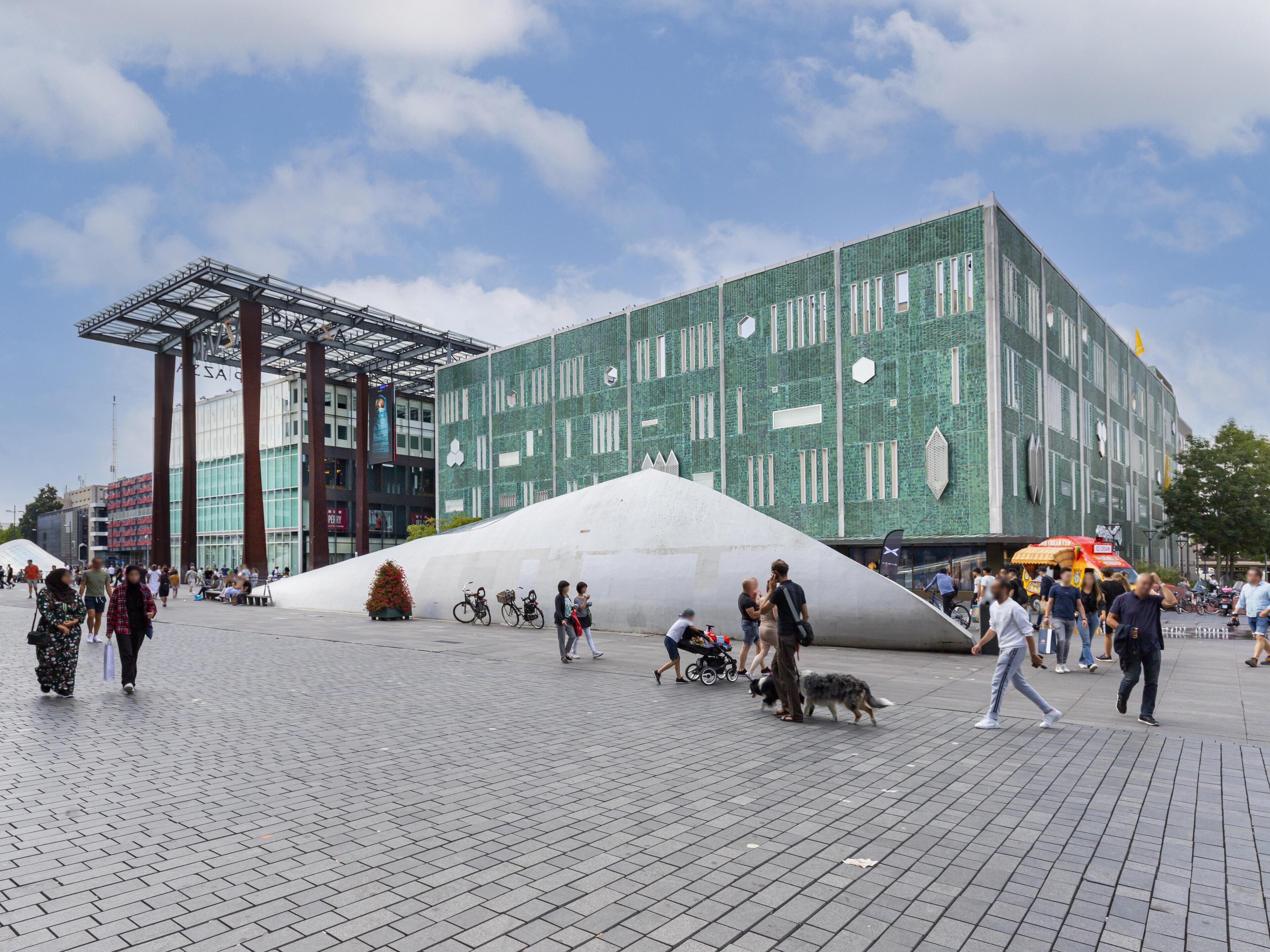 A public square with a modern building featuring a green facade and a large white sculpture.