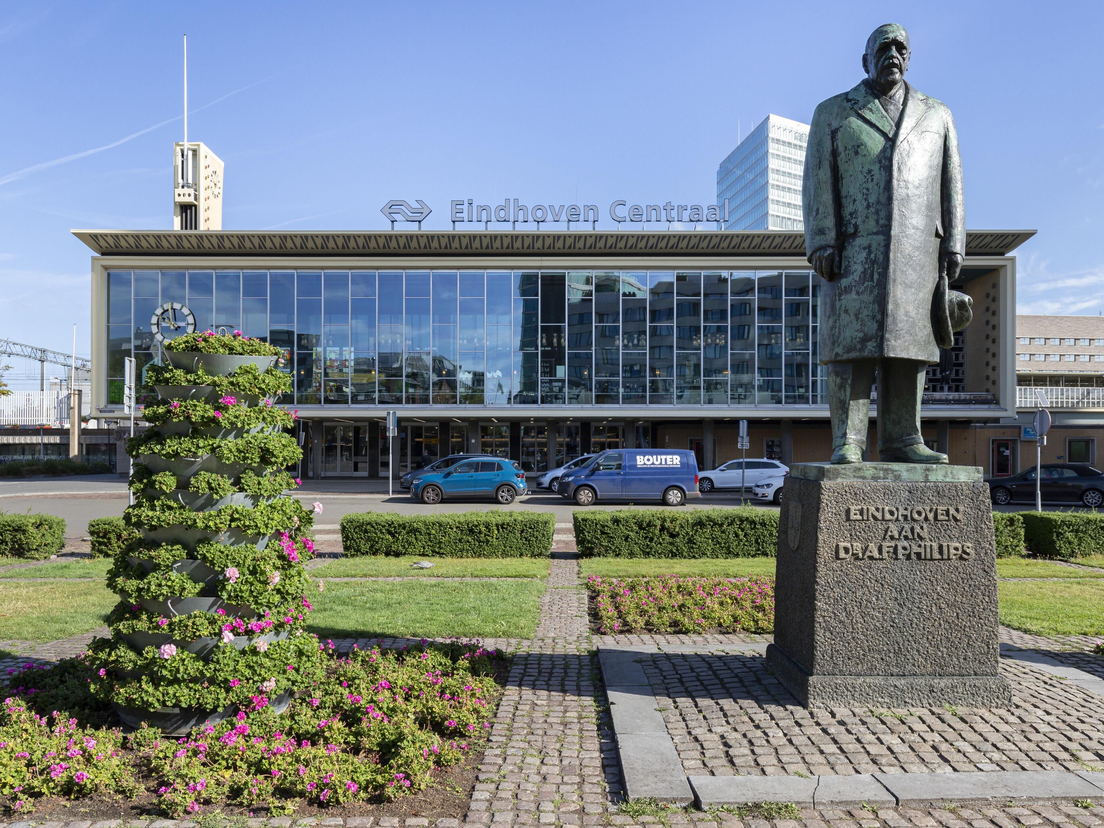 Eine Statue eines Mannes im Anzug, ein Gebäude mit der Beschriftung "Eindhoven Centraal" und ein blauer Transporter mit dem Schriftzug "BOUTER".
