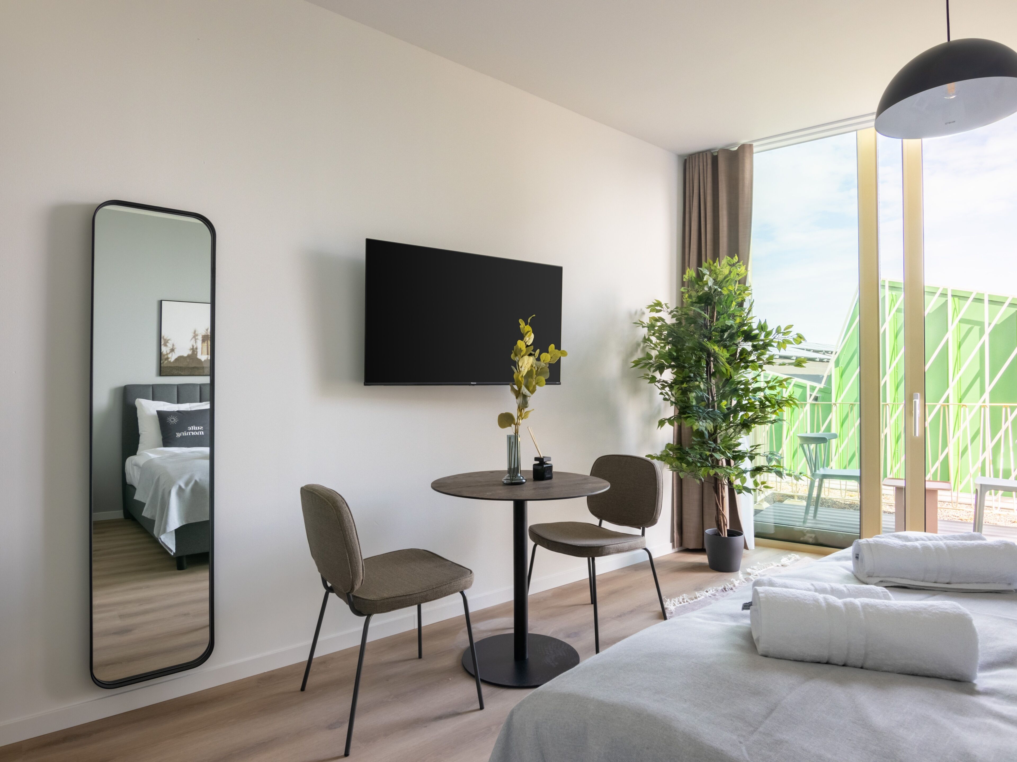 A modern hotel room with a bed, a round table with chairs, a large mirror, a flat-screen TV, and a potted plant.