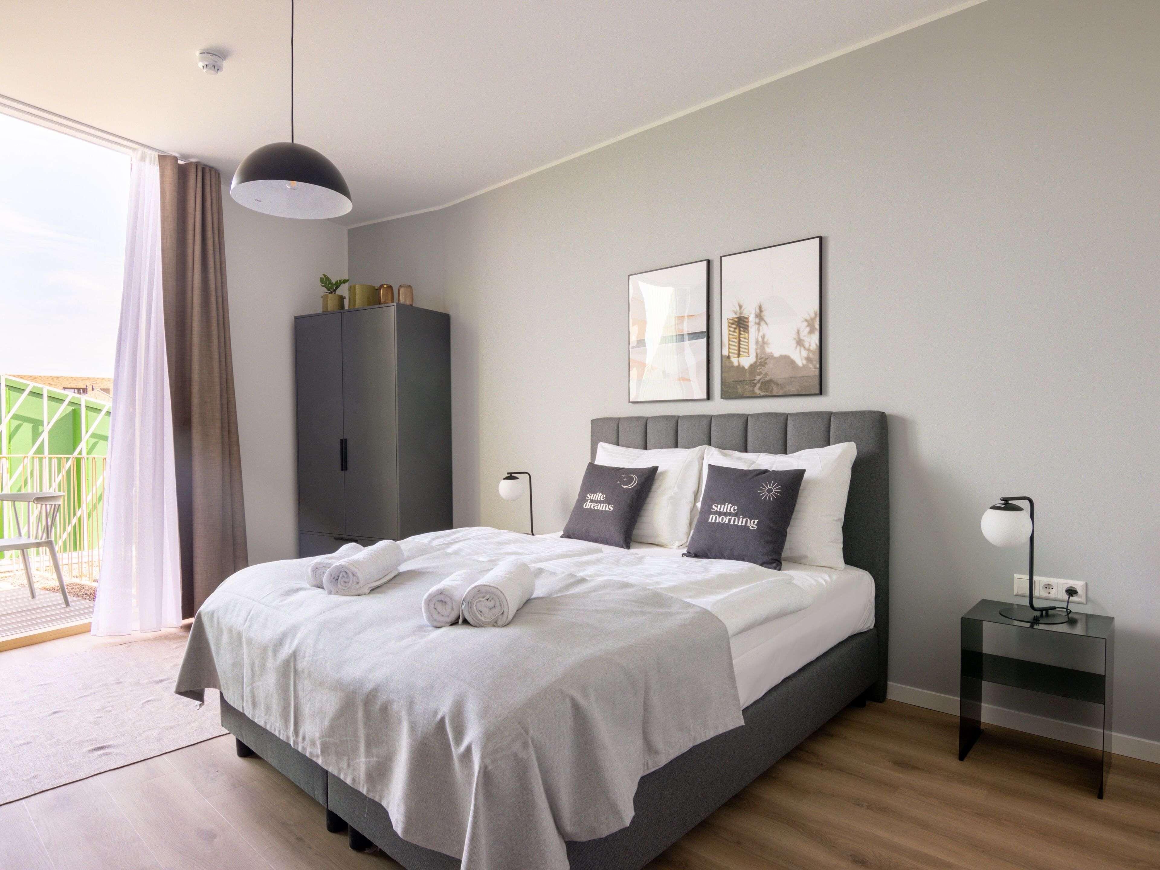 Un dormitorio moderno con cama, cabecero gris, dos mesitas de noche, un armario y un balcón.