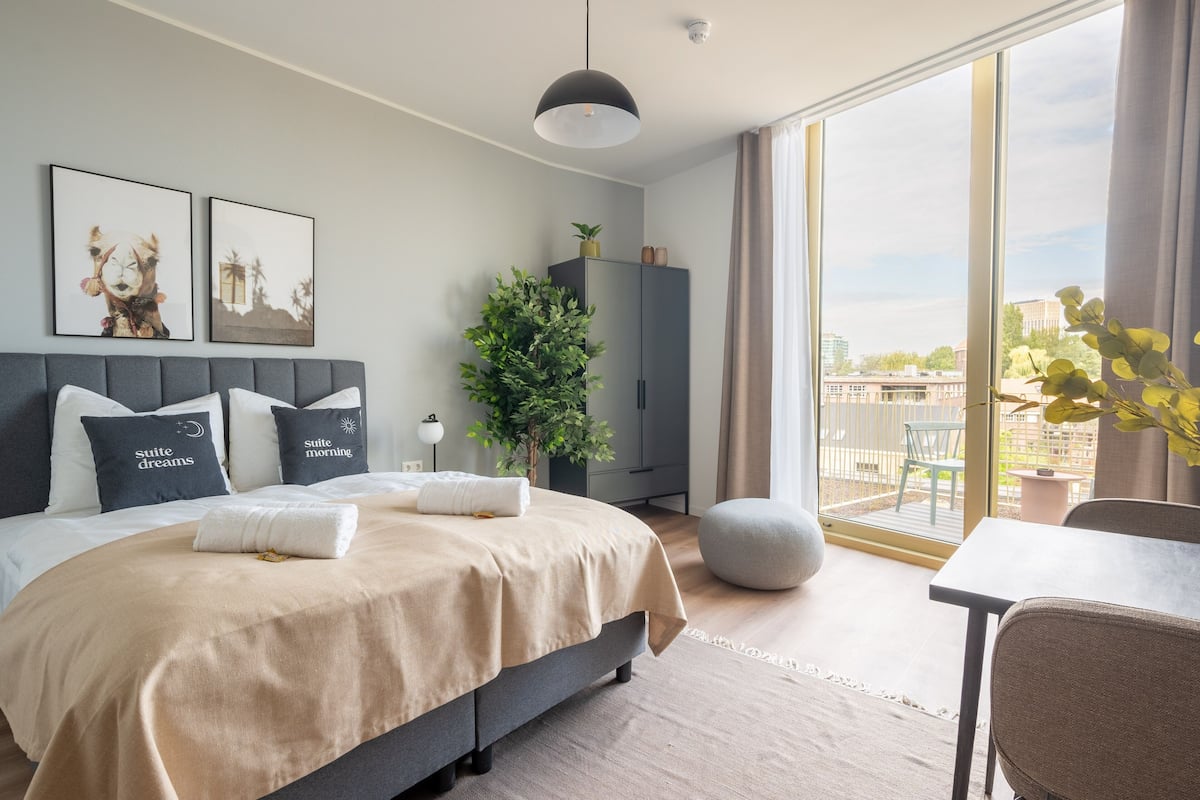 Een moderne slaapkamer met een groot bed, een bureau, een plant in pot en uitzicht op de stad door grote ramen.
