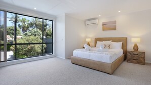 A spacious bedroom with a large window, a bed with white linens, two bedside tables with lamps, and an air conditioning unit on the wall.