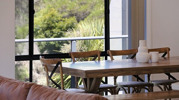 A dining area with a wooden table and chairs, a white vase, and a view of greenery through large windows.