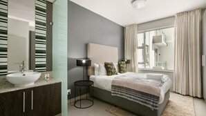 Ein modernes Schlafzimmer mit einem großen Bett, einem Nachttisch mit Lampe, einem Badezimmer mit Waschbecken und Spiegel sowie einem Fenster mit Vorhängen.