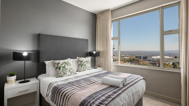Ein modernes Schlafzimmer mit großem Fenster, einem Bett mit gestreifter Bettwäsche, einem Nachttisch mit Lampe und Blick auf die Stadtlandschaft.