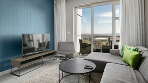 Ein modernes Wohnzimmer mit Flachbildfernseher, einem grauen Sofa, einem runden Couchtisch und einem großen Fenster mit Blick auf die Stadt.