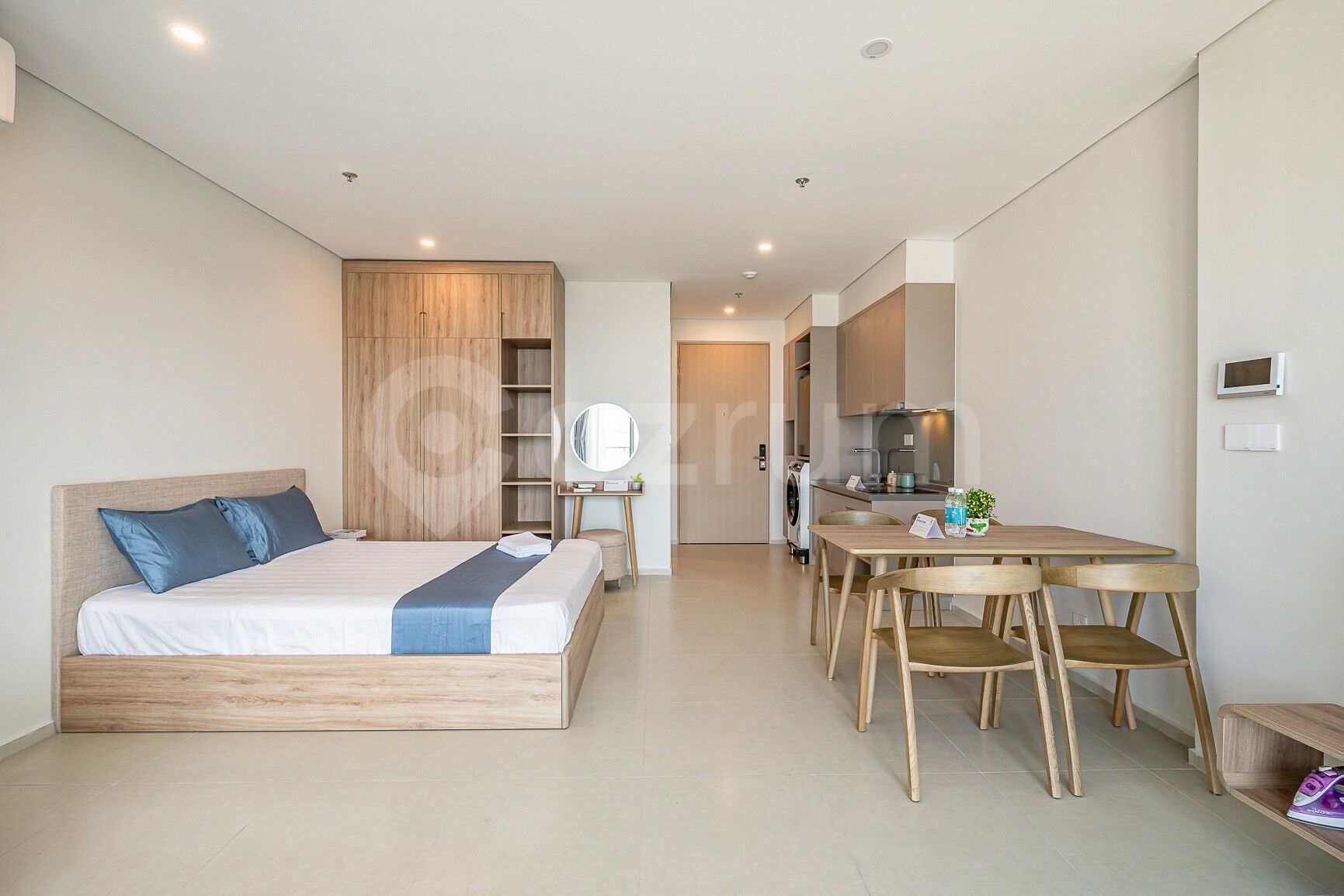 A modern bedroom with a wooden bed, a small desk, and a kitchenette in the background.