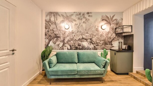 Una sala de estar con un sofá verde, un mural de follaje tropical y una pequeña zona de cocina con un refrigerador y un lavabo.