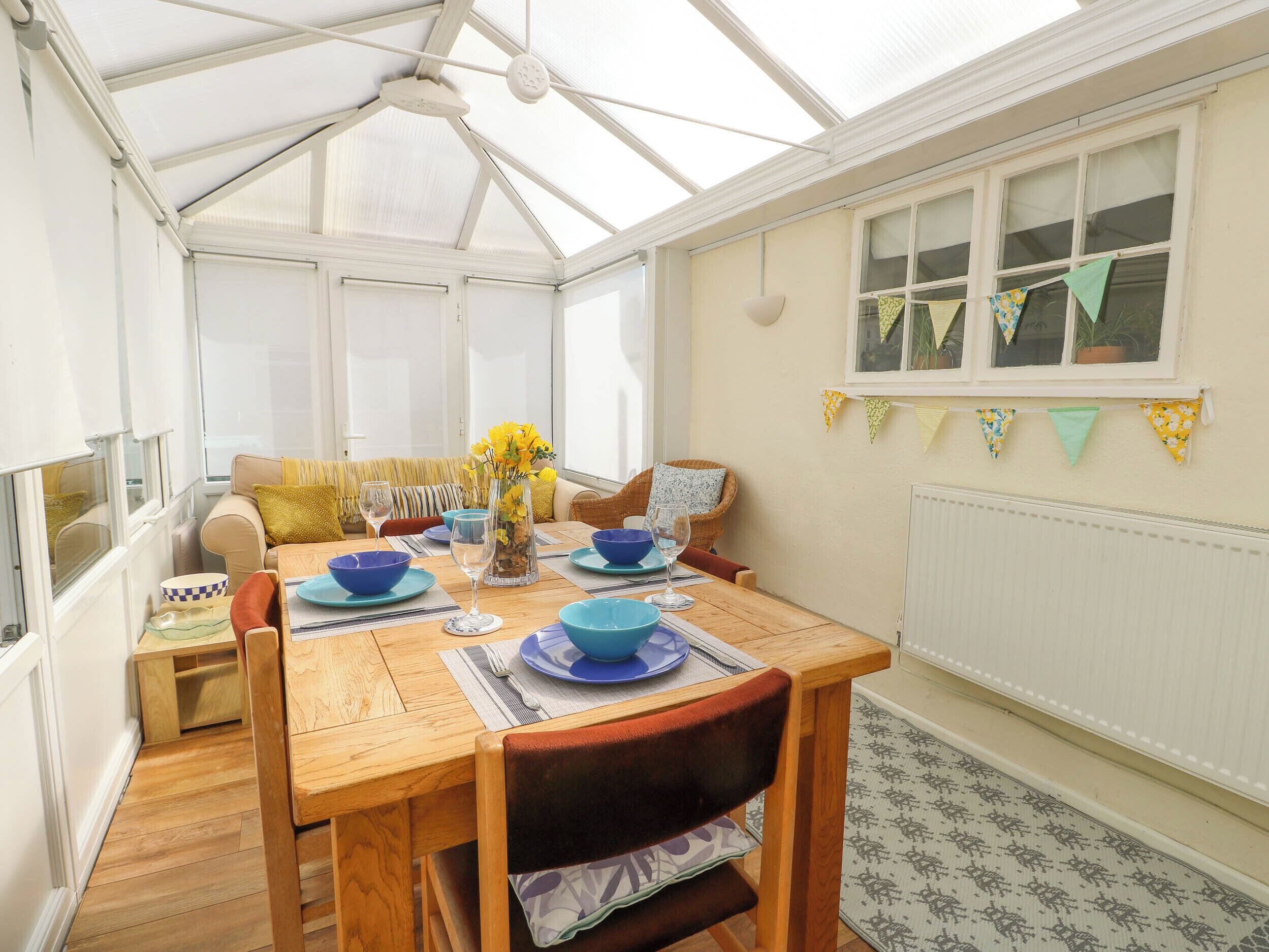 A conservatory with a wooden dining table set for four, surrounded by wicker chairs.