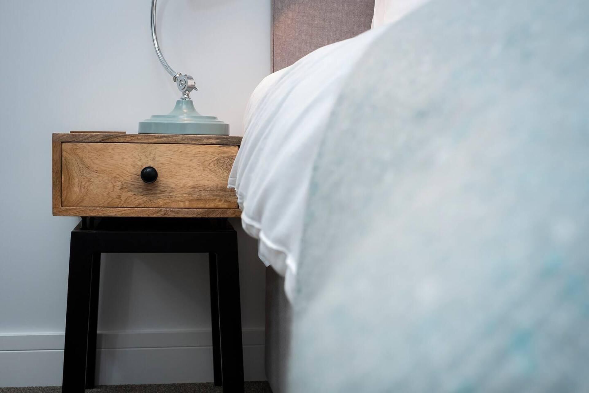 A wooden bedside table with a lamp and a drawer, next to a bed with white bedding.