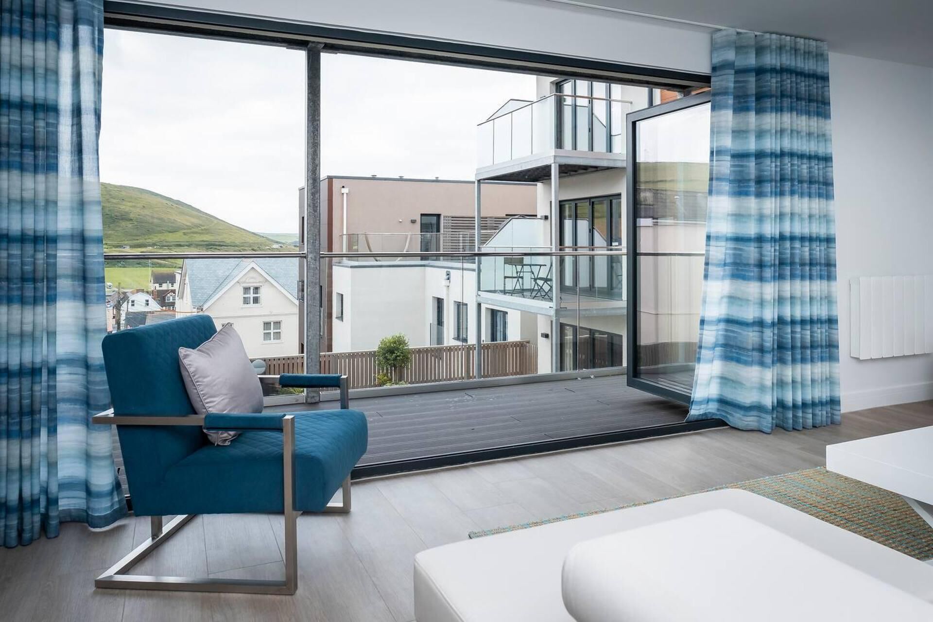 A modern living room with a blue armchair, a white sofa, and a large window offering a view of a multi-story building and a hill.