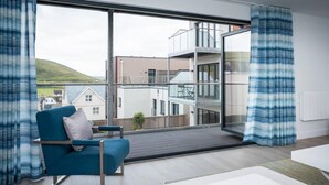 A modern living room with a blue armchair, a white sofa, and a large window offering a view of a multi-story building and a hill. - This Apartment is No Longer Available (Woolacombe)