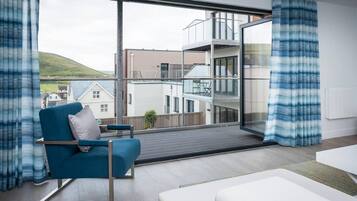 A modern living room with a blue armchair, a white sofa, and a large window offering a view of a multi-story building and a hill.