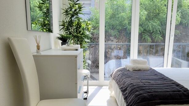 A modern bedroom with a white bed, a white chair, a desk, and a large window offering a view of greenery outside. - Central and cozy- FREE PARKING (Lugano)