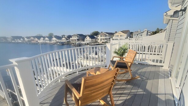 Een houten terras met twee schommelstoelen en een kleine tafel, met uitzicht op een jachthaven met boten en huizen op de achtergrond.