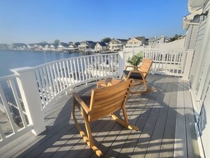 Um deck de madeira com duas cadeiras de balanço e uma pequena mesa, com vista para uma marina com barcos e casas ao fundo.