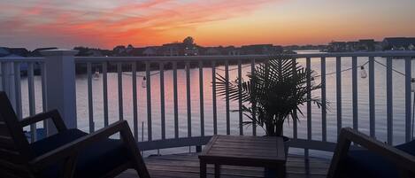 A wooden deck with a table and chairs, overlooking a body of water with a sunset in the background.