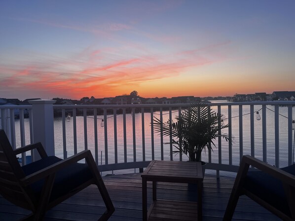 Um deck de madeira com uma mesa e cadeiras, com vista para um corpo de água e um pôr do sol ao fundo.