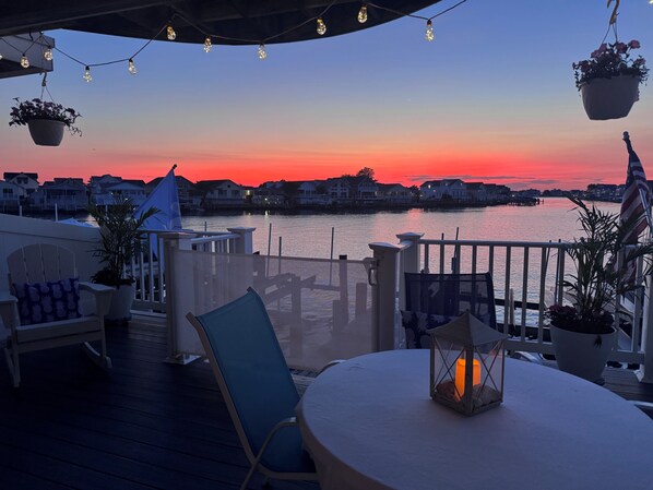 A deck with chairs, a table, hanging plants, and a view of the water and houses at sunset.