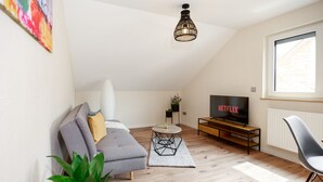 A modern living room with a grey sofa, a coffee table, a TV displaying Netflix, a plant, and a window with natural light.