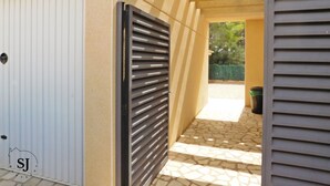 A gated entrance with a metal gate and a white door, leading to a covered outdoor area with a tiled floor and a view of greenery. - Pleasant villa with pool (A) (Ametlla de Mar (Les Tres Cales))
