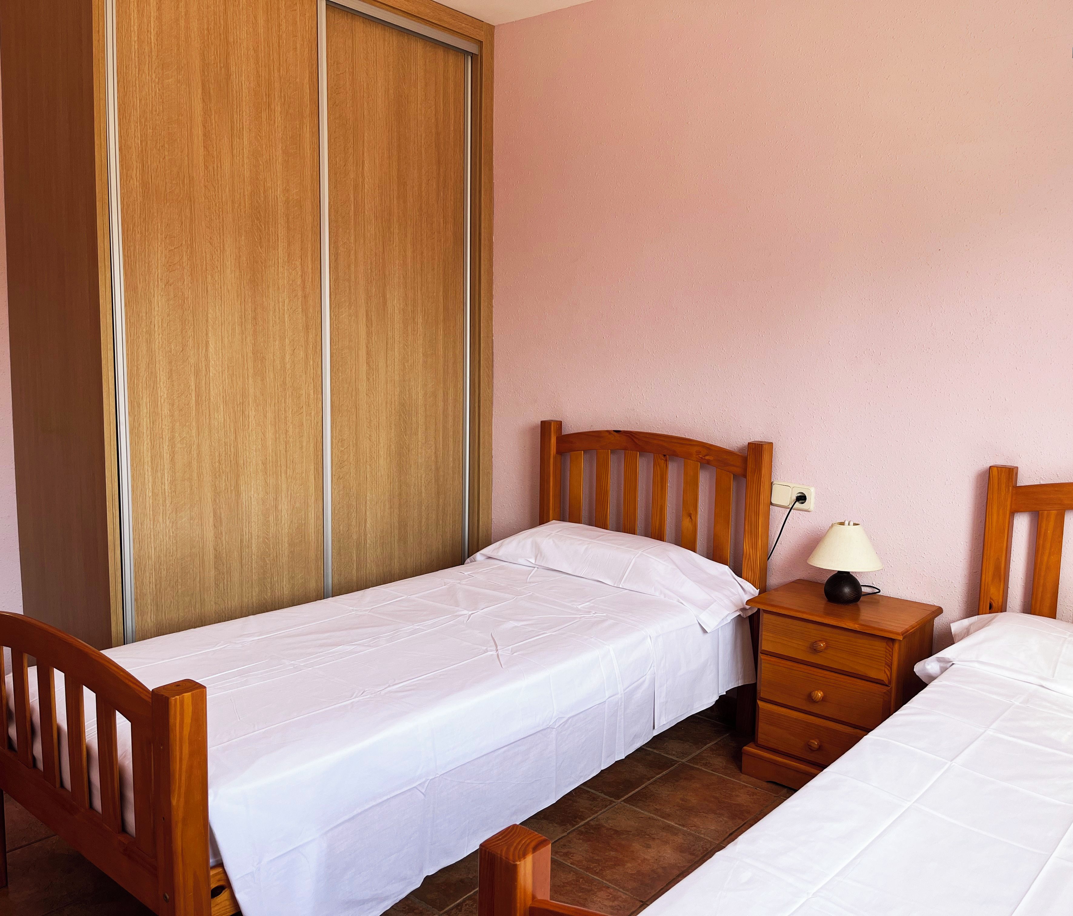Un dormitorio con dos camas individuales, un armario de madera, una mesita de noche con lámpara y un logotipo en la pared.