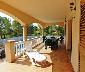 Une terrasse extérieure couverte, avec un fauteuil de relaxation blanc, une table avec des chaises et une vue sur les arbres et une clôture.