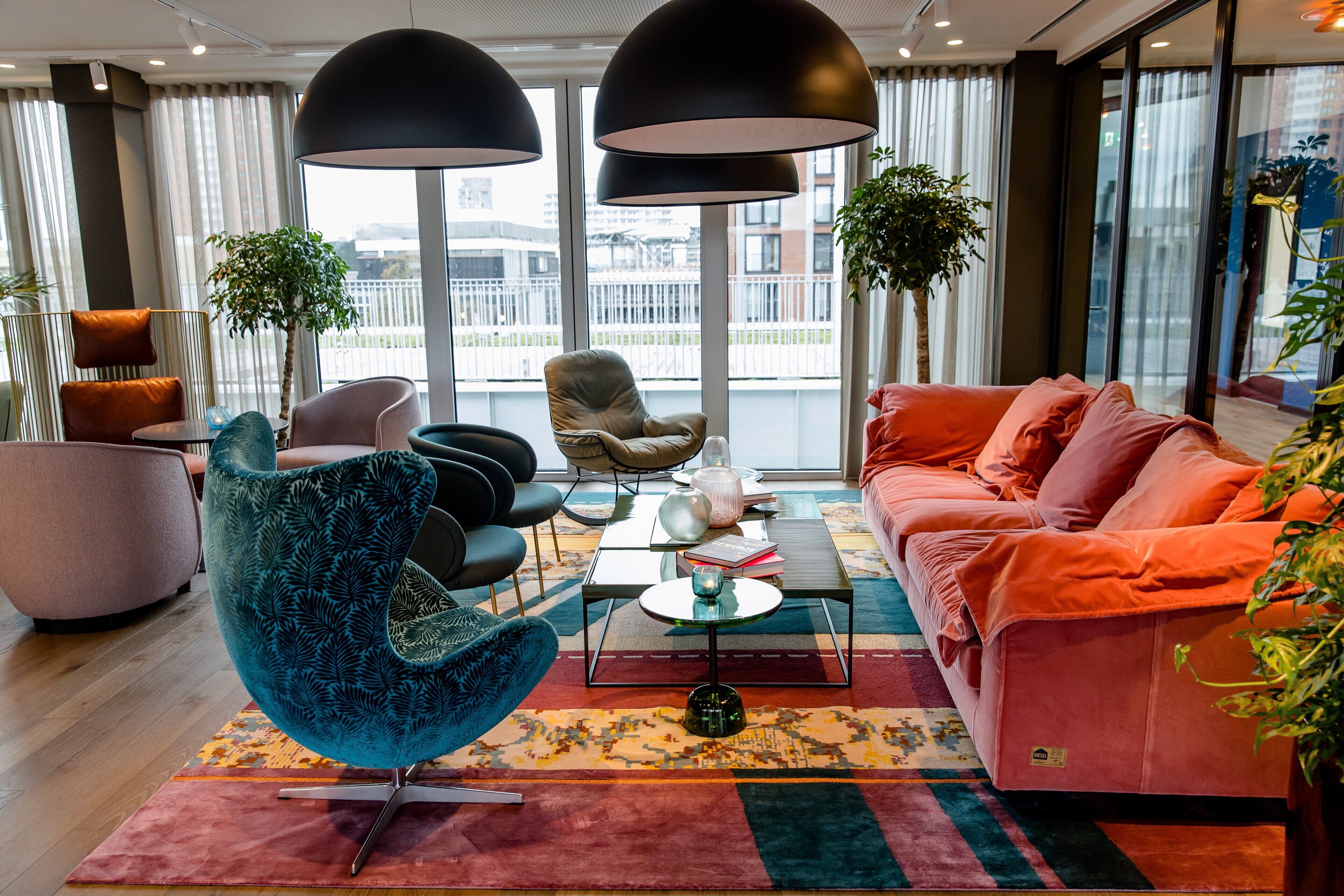 Una sala de estar moderna con un sillón verde azulado, un sofá rosa y una mesa de centro. Hay dos grandes lámparas colgantes negras y una vista de la ciudad a través de amplias ventanas.