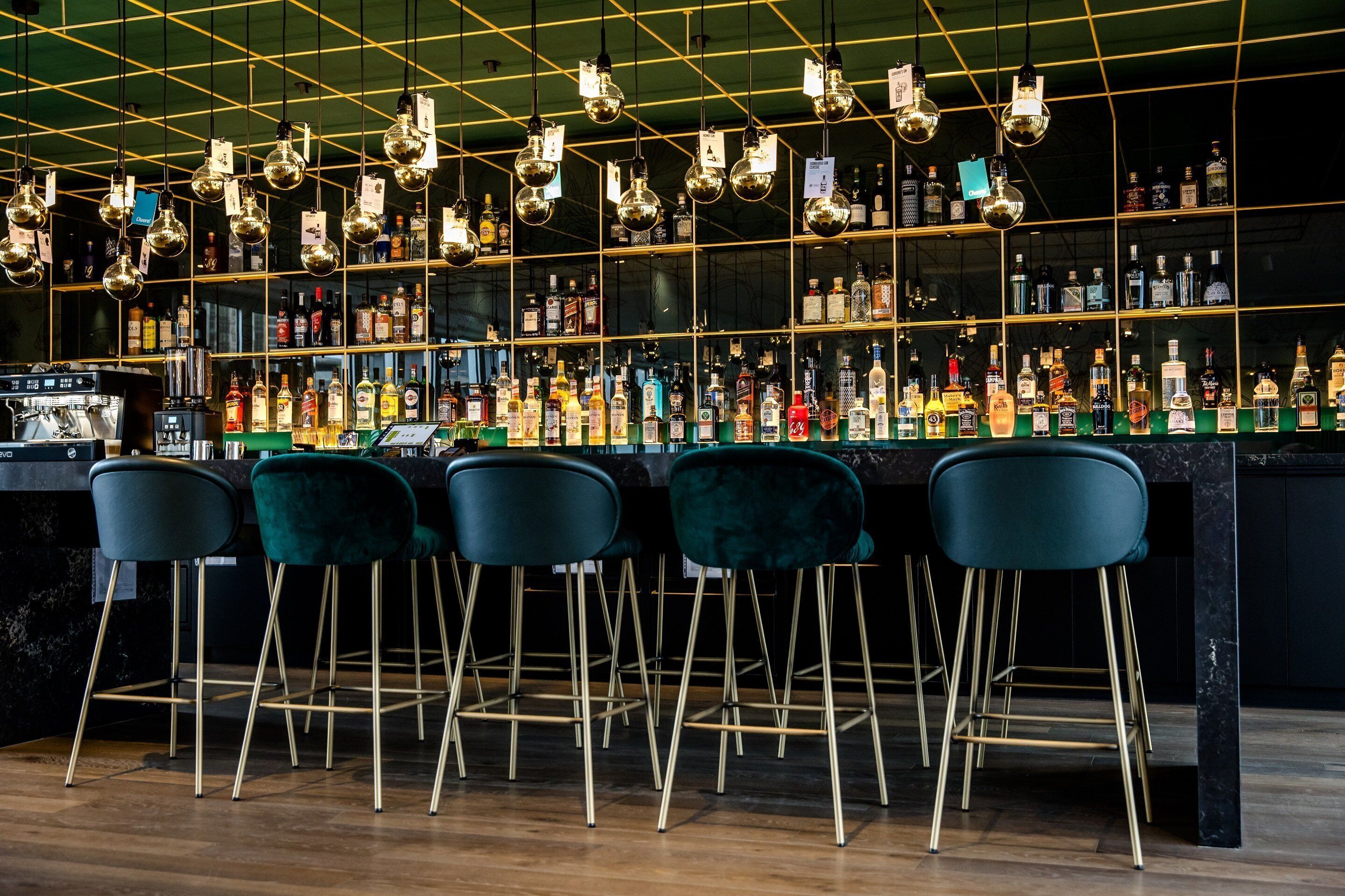 A bar area with multiple liquor bottles, green bar stools, and hanging light fixtures.