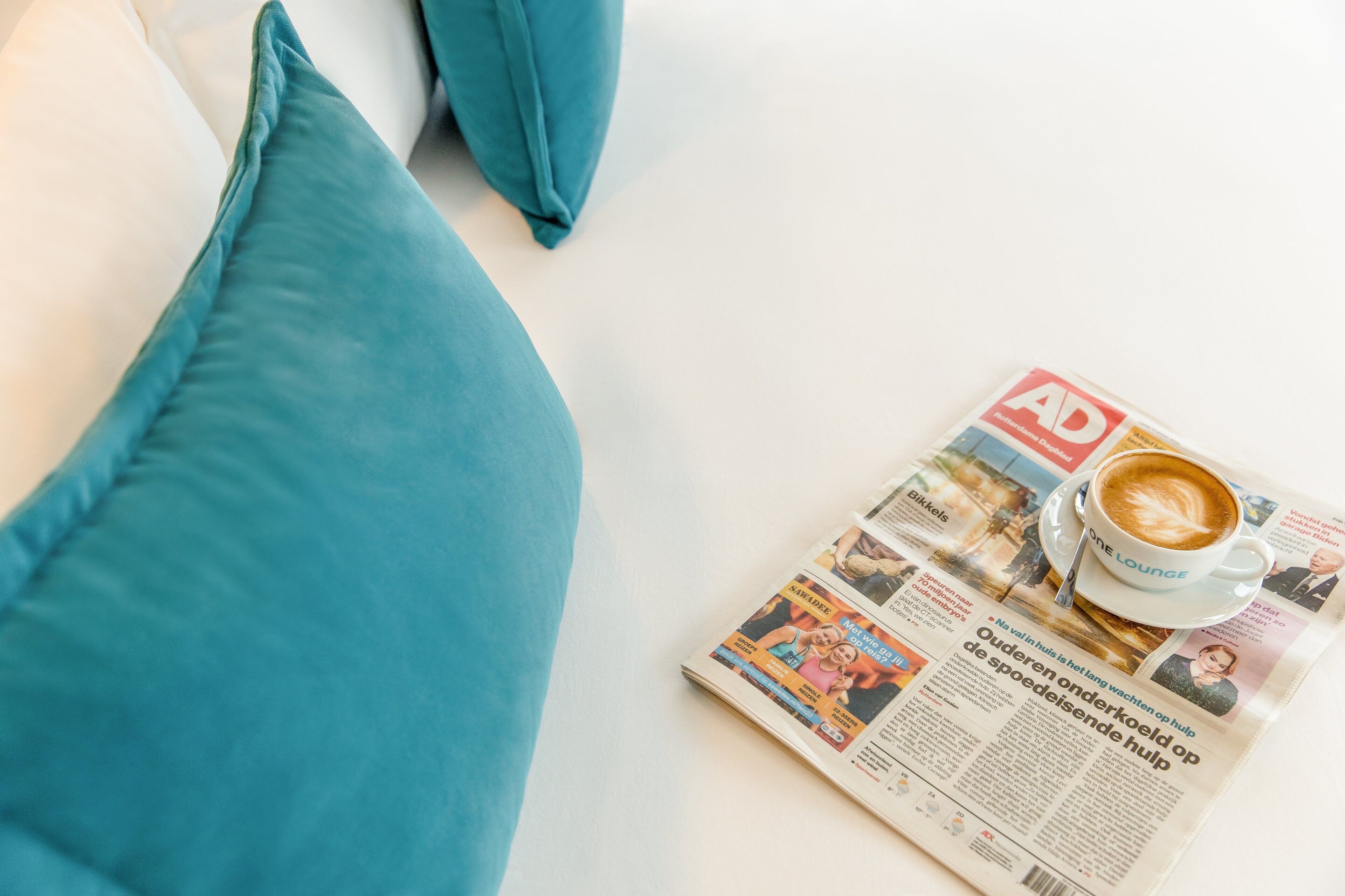 A newspaper with a coffee cup on a table, surrounded by teal pillows.