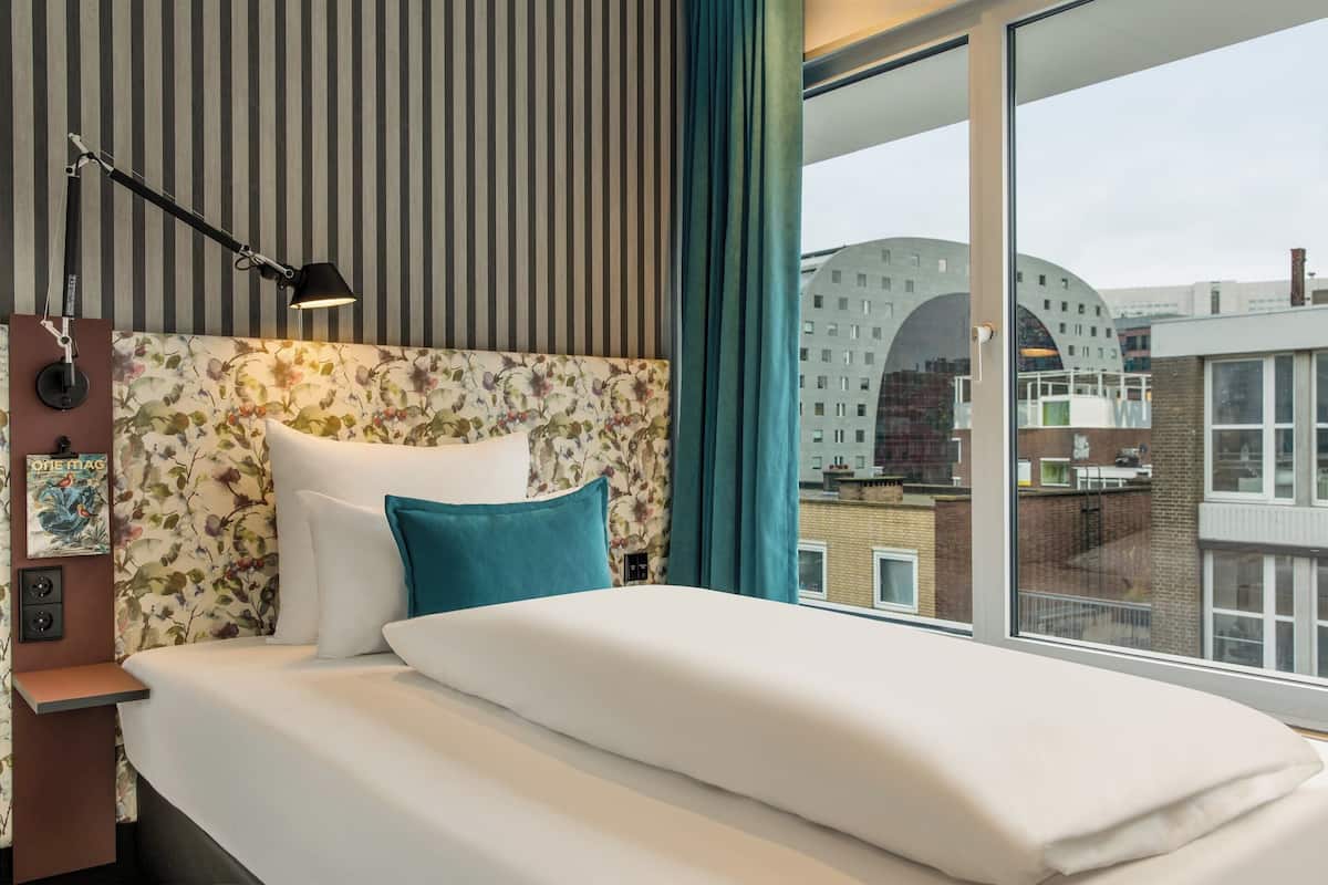 Habitación de hotel con una cama grande, una mesita de noche con lámpara, cabecero floral y vistas a la ciudad a través de un ventanal amplio.