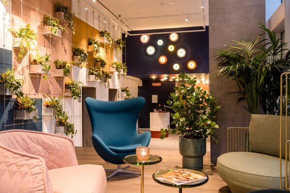 A modern interior with a blue armchair, a pink sofa, and a green armchair. There are multiple potted plants and a large black wall with circular light fixtures.