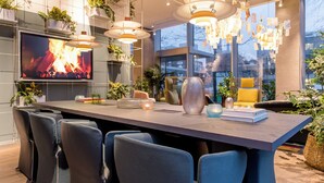 A modern dining area with a long table, multiple chairs, pendant lights, and potted plants.