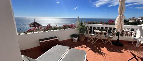 A balcony with a view of the sea, a table with chairs, a bench, and a potted plant.