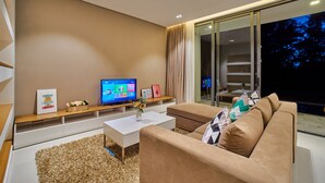 Ein modernes Wohnzimmer mit Flachbildfernseher, weißem Couchtisch, beiger Couch und großem Fenster mit Vorhängen.