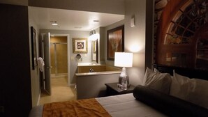 Een hotelkamer met een bed, een nachtkastje met een lamp, een badkamer met een spiegel en een schilderij aan de muur.