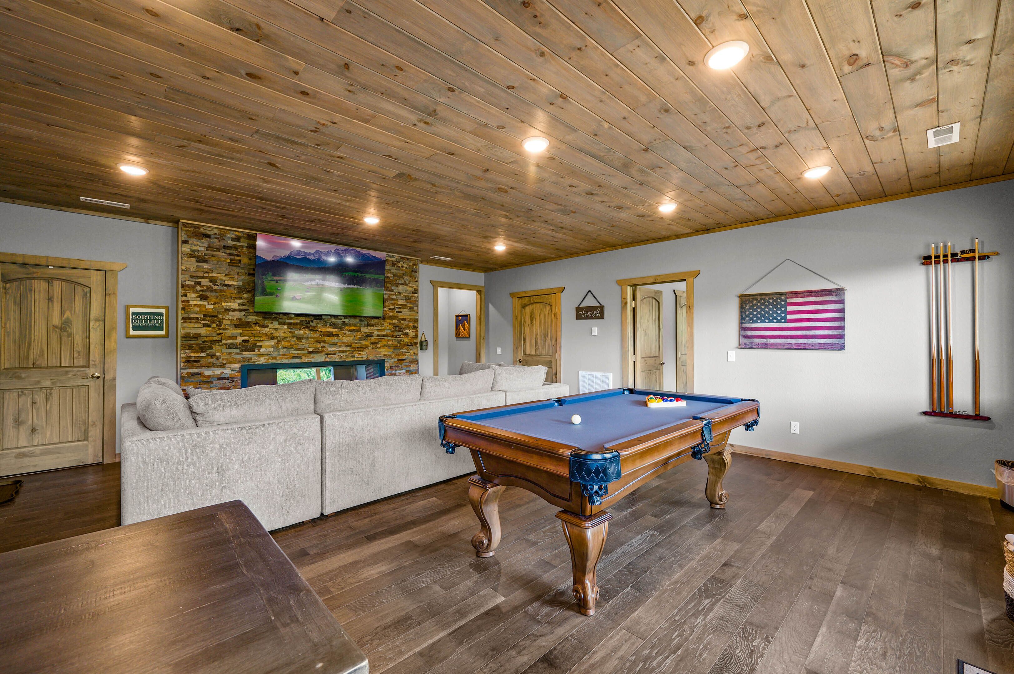 A pool table in a room with wooden flooring, a stone wall, a television, a sofa, and an American flag.