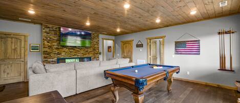 A pool table in a room with wooden flooring, a stone wall, a television, a sofa, and an American flag.