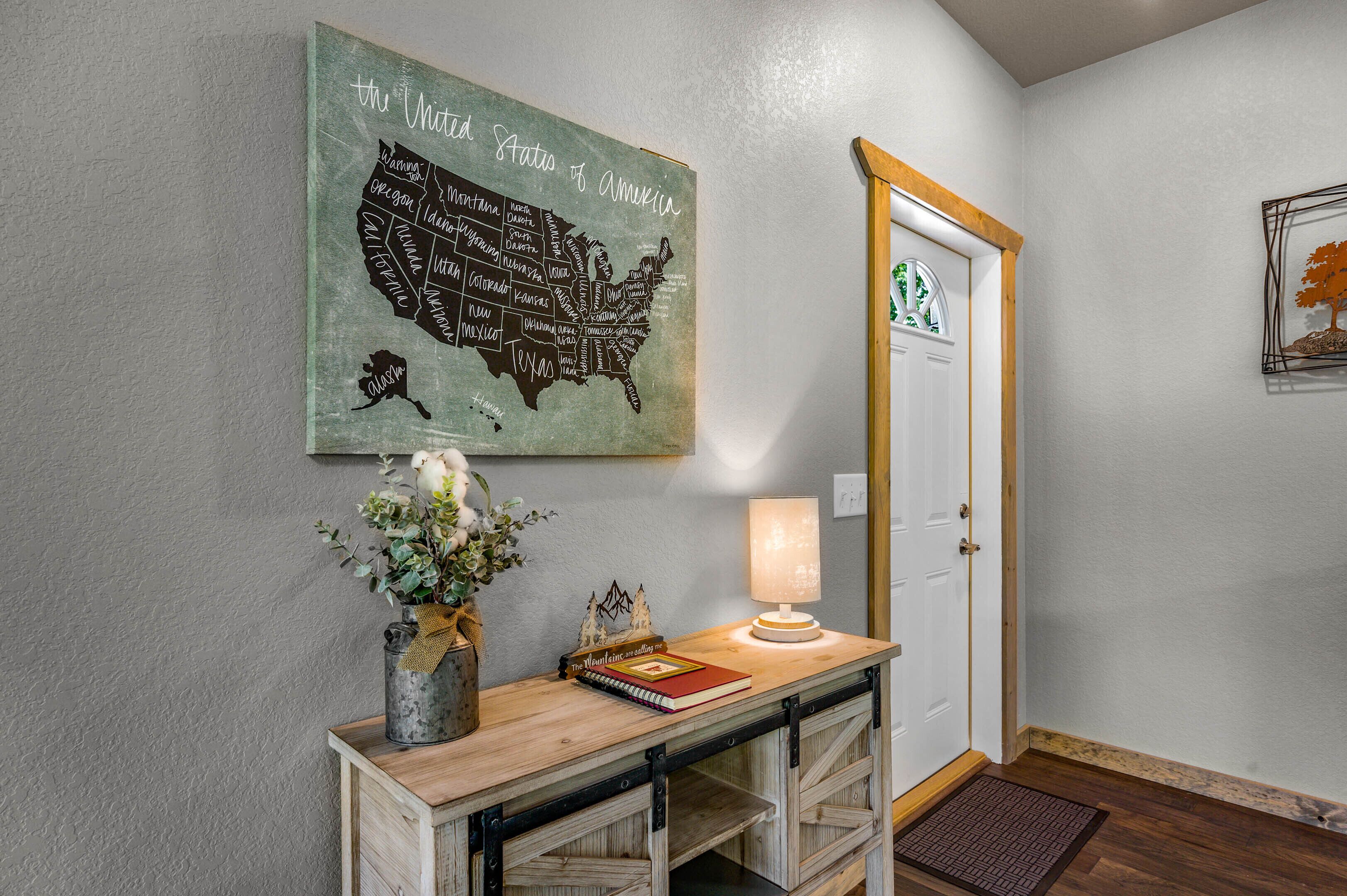 Un couloir orné d’une carte des États-Unis, une console en bois sur laquelle repose une lampe, un vase rempli de fleurs et une porte avec une enseigne verte.