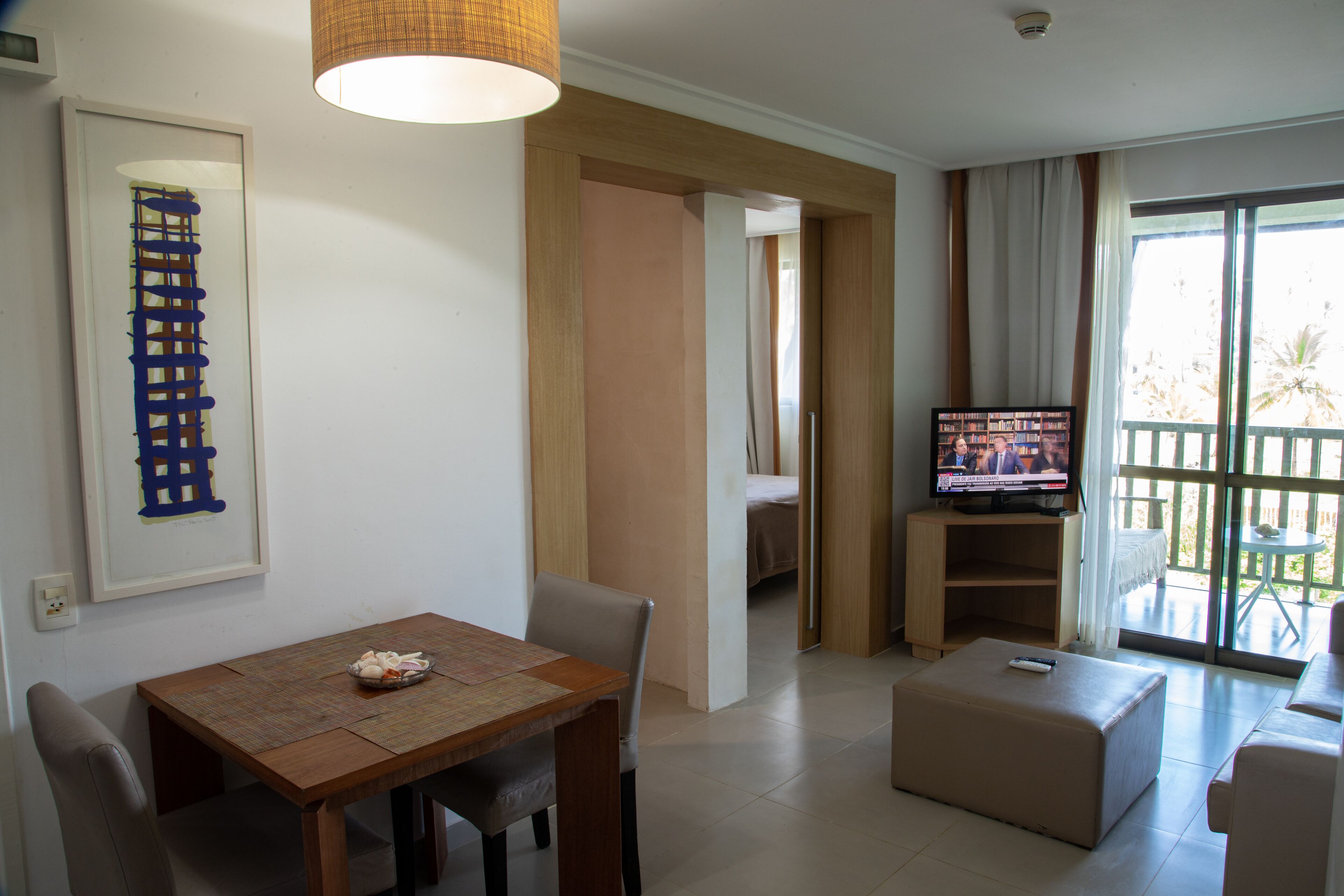 A living room with a wooden dining table, a sofa, a TV, and a balcony with a view.