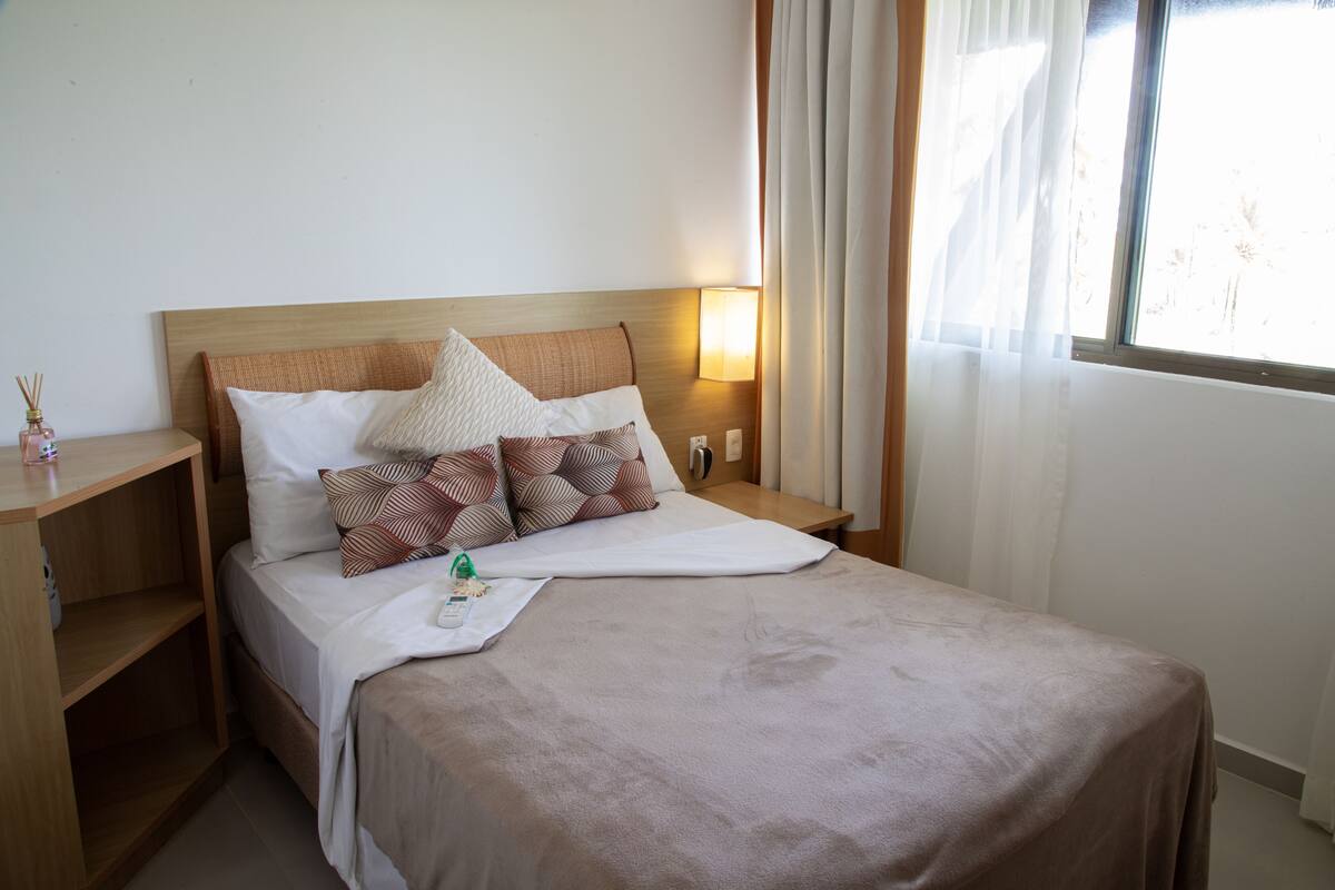 A neatly made bed with a wooden headboard, a bedside table with a lamp, and a window with curtains.