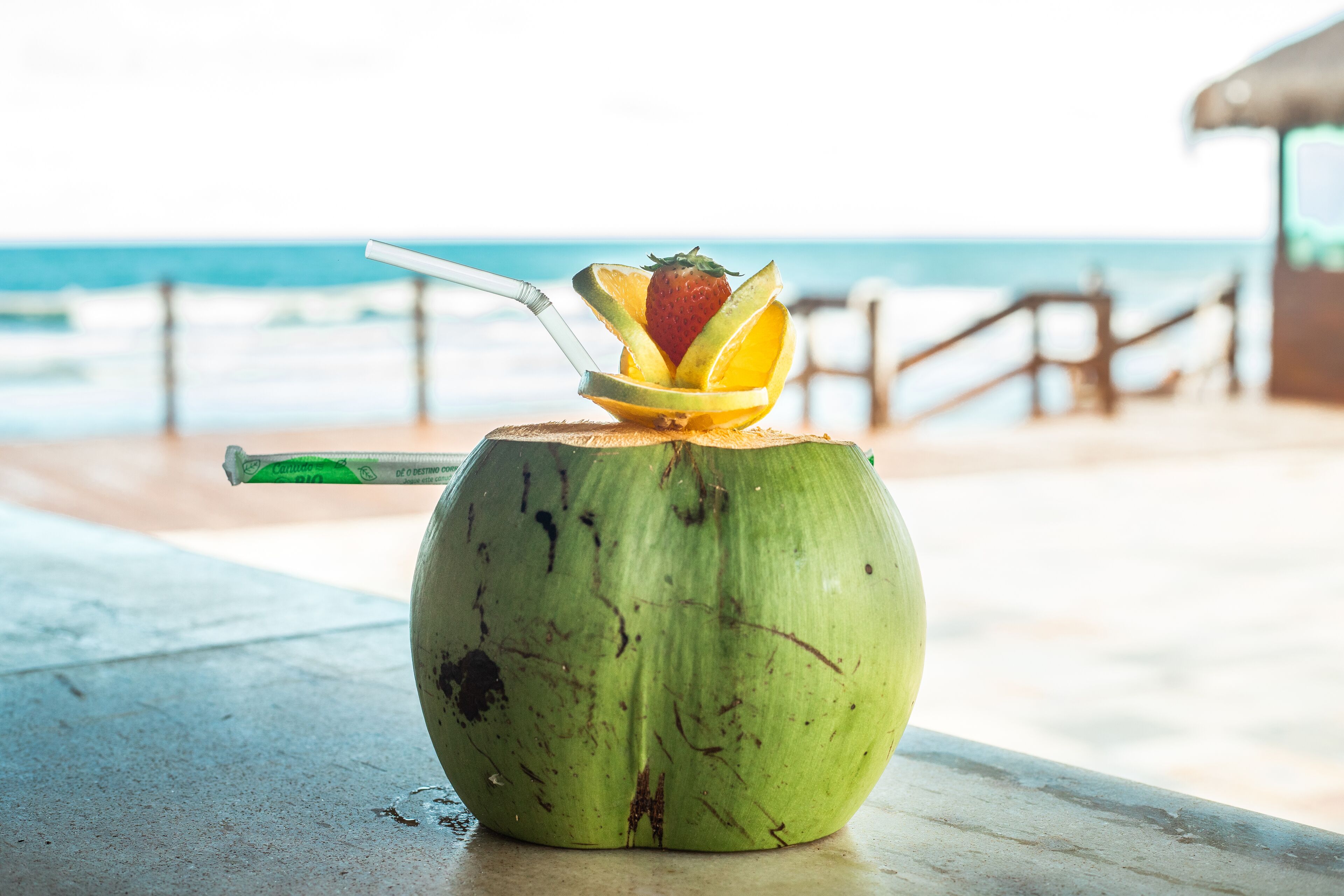 A green coconut with a straw and a fruit garnish on a table by the beach.