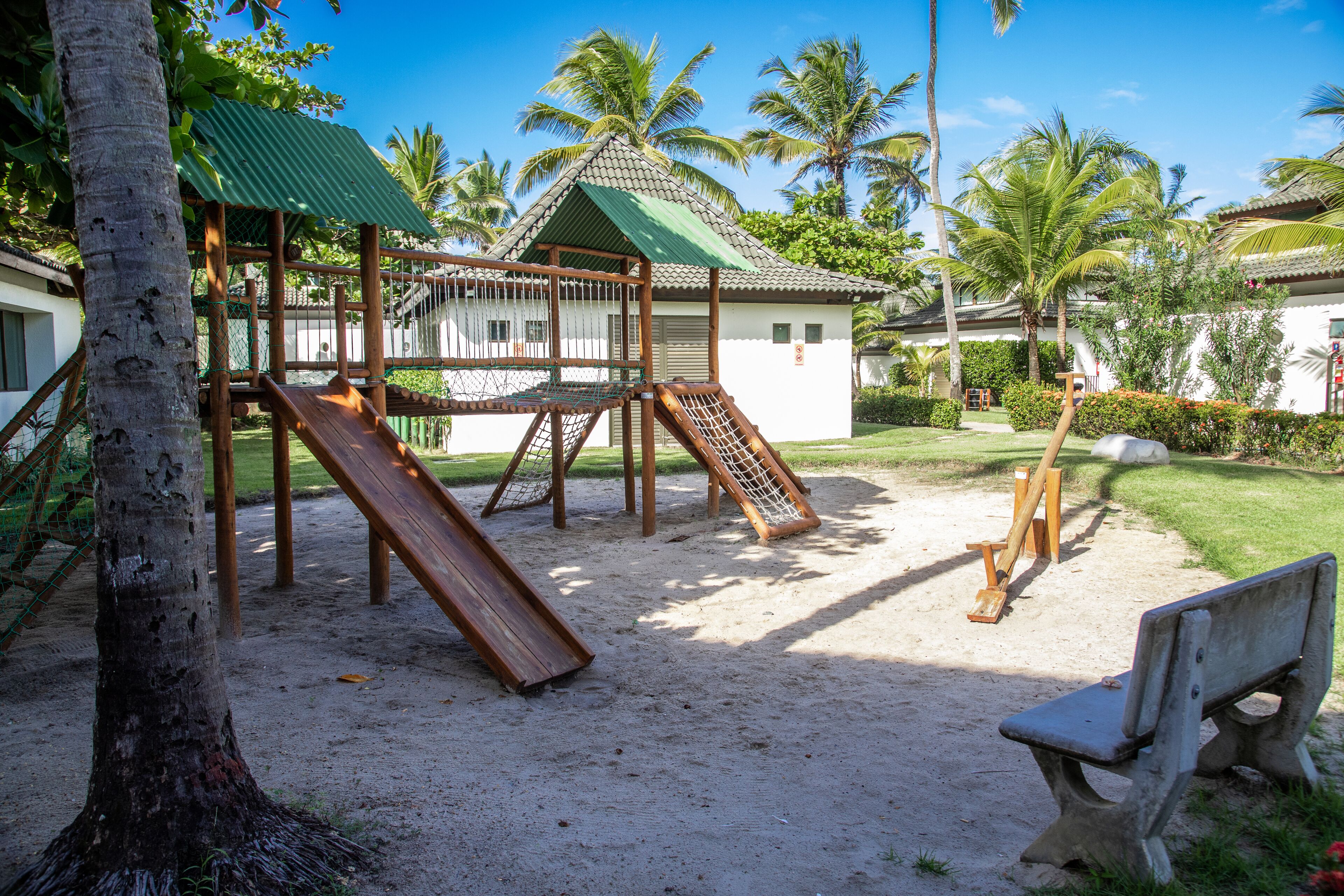 Un parque infantil con toboganes y estructuras para escalar, rodeado de palmeras y una zona de arena.