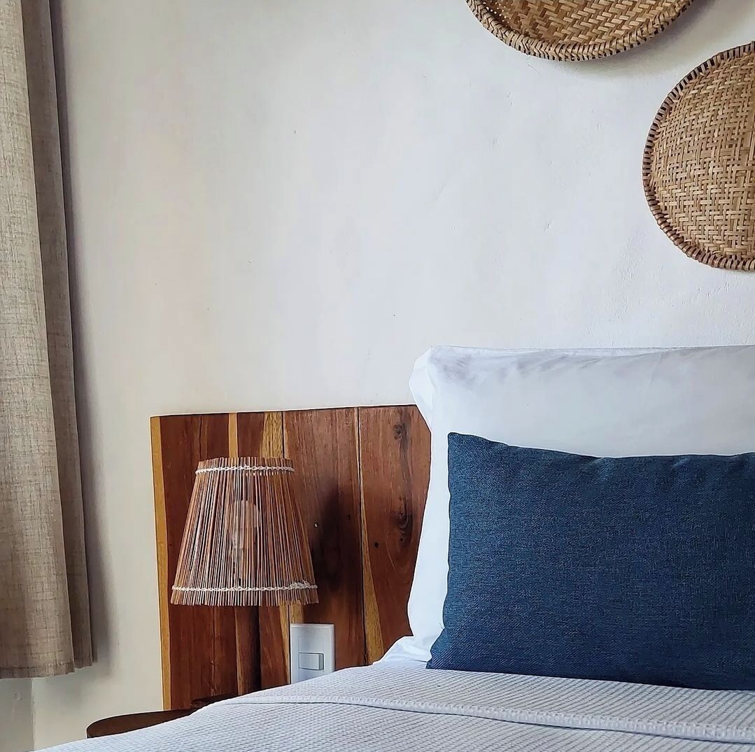Una cama bien hecha con almohada blanca y azul, cabecera de madera y lámpara tejida sobre una mesita de noche.
