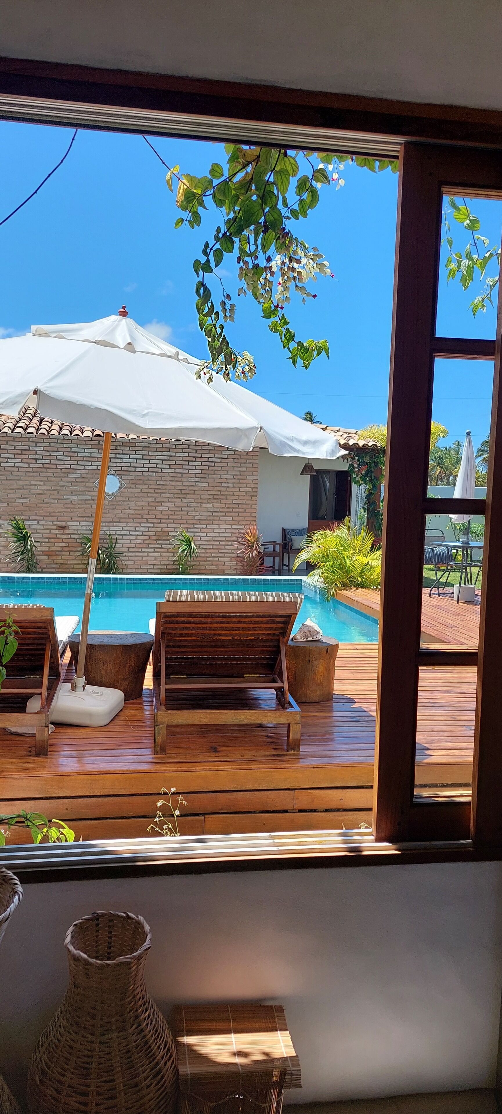 Una terraza de madera con sillones de descanso y piscina, una sombrilla blanca y un muro de ladrillo.